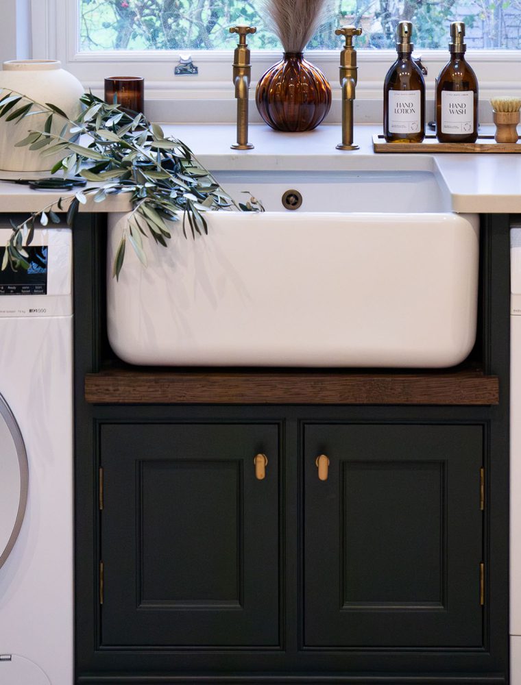 A bespoke bootroom in Downpipe. Two Siemens washing and drying appliances, set within bespoke cabinetry, command attention. So too, the Belfast sink set into the white worktop above the appliances. On the right side of the worktop, two wicker baskets are set against the wall. Beside the sink, hand wash and lotion stand opposite the small white vase next to the sink’s edge. On the right side, and on the from the vase, two baskets containing hand towels and clothes pegs stand next to a tray with incense on it.