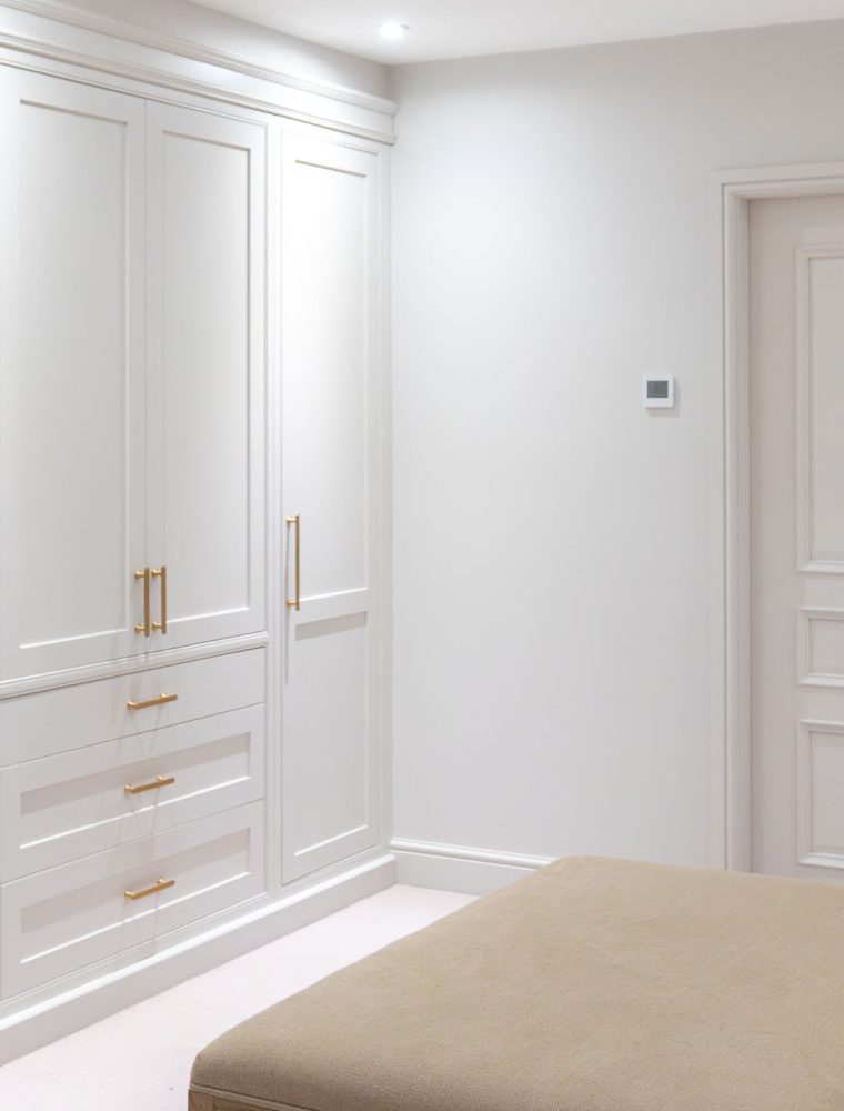 A bespoke dressing room in French Grey. A floor-to-ceiling in frame wardrobe stands against the far wall on a right angle from the door. In the middle of the floor stands an upholstered bench. Both door and wardrobe have Corston Architectural handles.