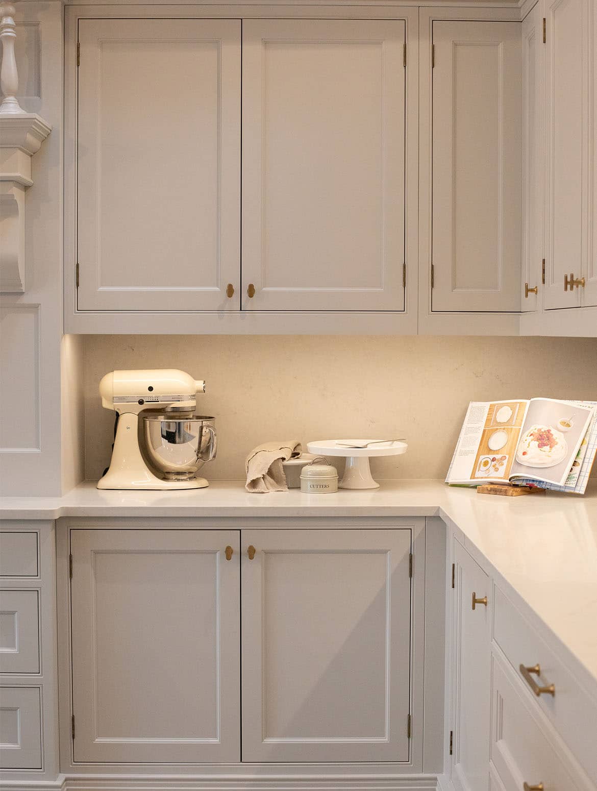 A traditional shaker kitchen in French Grey.  Bespoke cabinets above and below sit at right angle join to the kitchen wall. A standing food mixer and cake pedestal sit next a propped-up baking recipe book. A ornate mantel is just visible as well. 