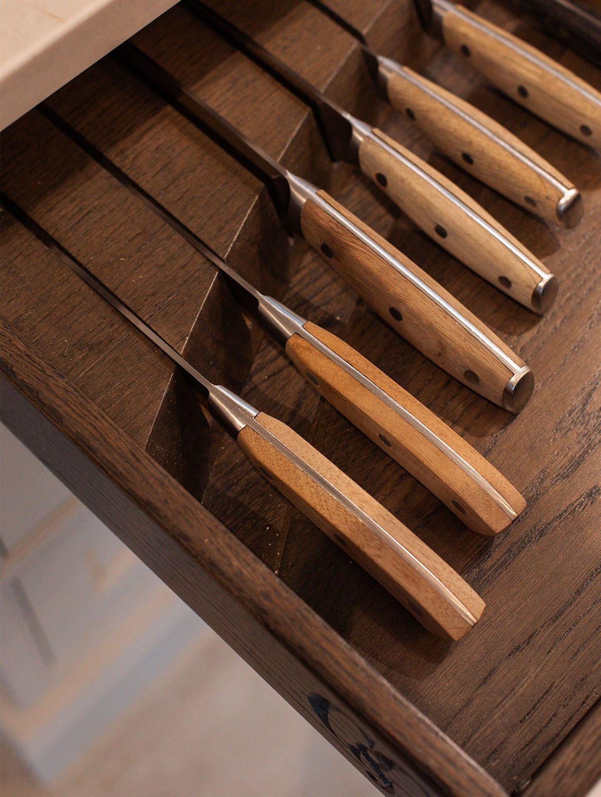 A traditional shaker kitchen in French Grey. A selection of knives with wooden handles in a bespoke island drawer run from largest to smallest. The Cooks & Company logo is embossed into the side of the drawer. 