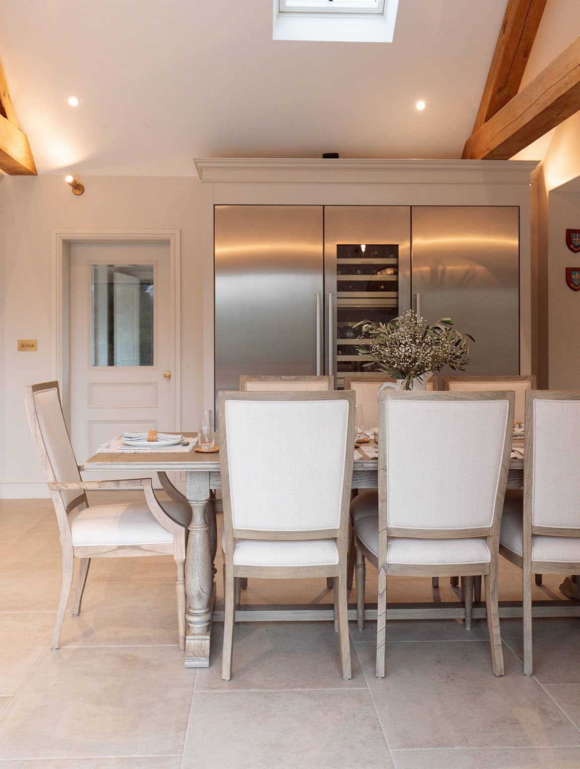 A traditional shaker kitchen in French Grey. A dining table set for a meal sits before a Liebherr monolith refrigeration bank set within bespoke cabinetry. Above, well-lit beams oversee the kitchen. 