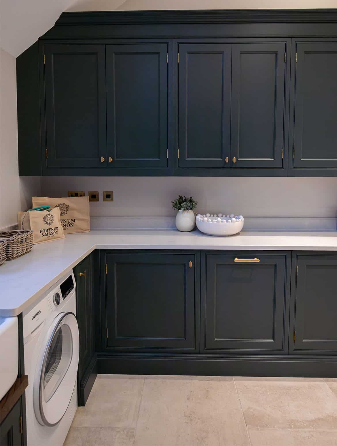 A bespoke bootroom in Downpipe. The left dog-leg of the bespoke cabinetry shows off a Siemens washing machine below, while the white worktop above contrasts the Armac Martin handles on the cupboard doors. Two Fortnum & Mason bags sit in the corner and on from them them two ceramic vases catch the eye. 