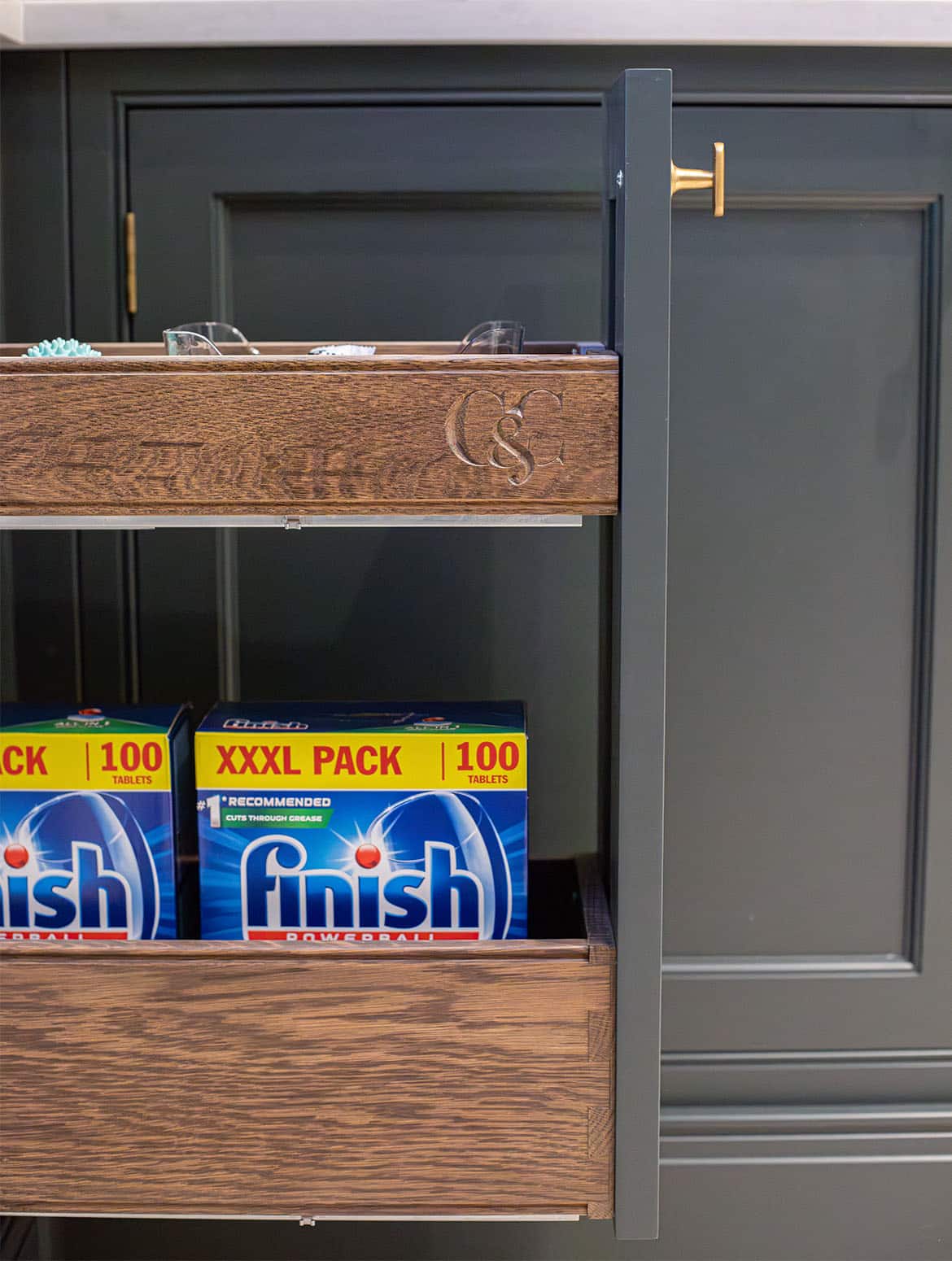 A bespoke bootroom in Downpipe. A close in view of an open drawer with an Armac Martin handle on it. In contrast to the darker colour of the panelling, the light wood of the drawer has the C & C logo of Cooks & Company engraved into it. On the bottom drawer, two boxes of laundry detergent are visible.  