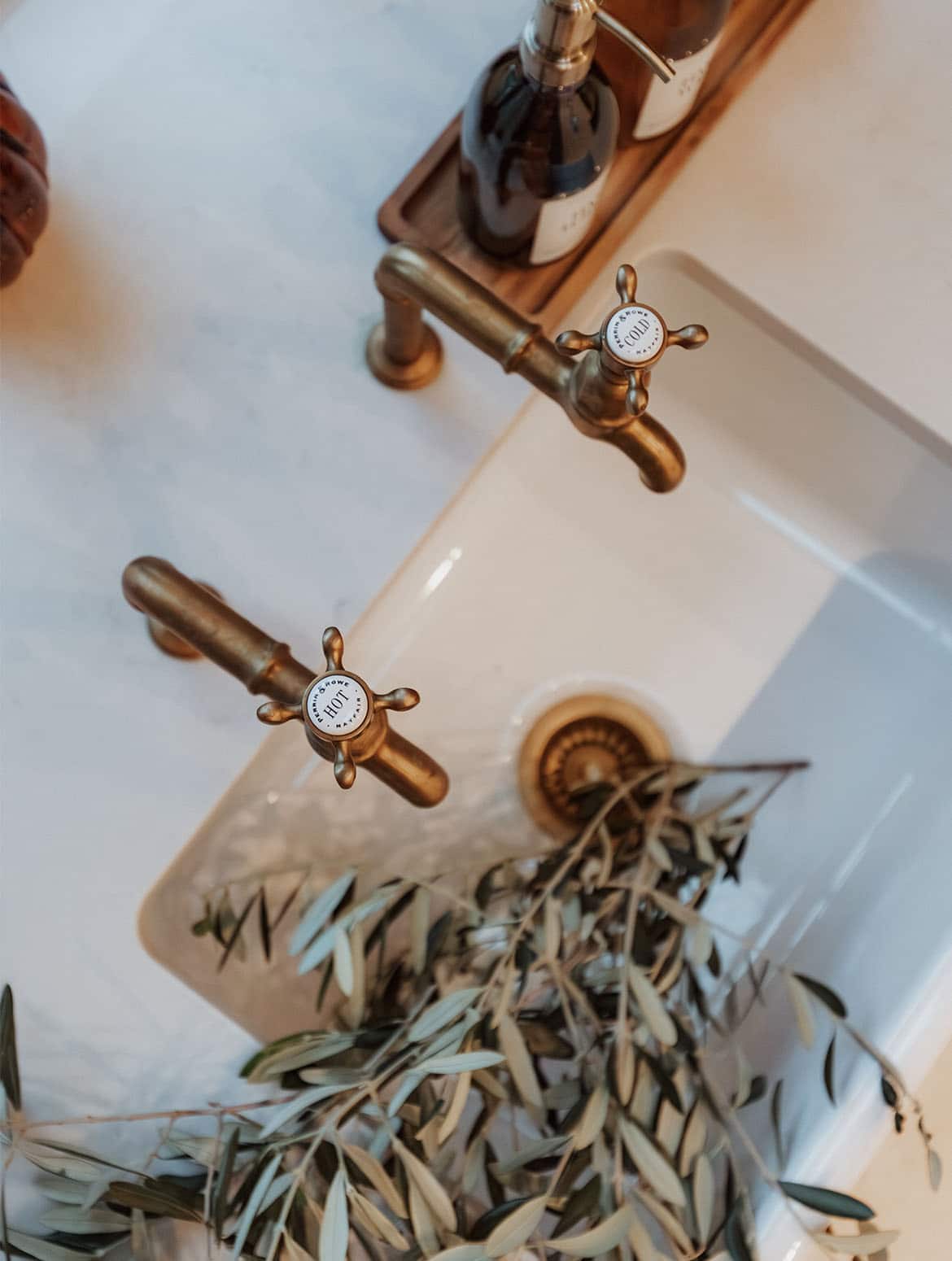 A bespoke bootroom in Downpipe. A bird’s-eye-view of the Belfast sink with its pair of burnished bronze Perrin and Rowe bibcock taps and bay leaves in the sink. There’s a dish with hand soap and lotion dispensers next to the cold tap. 