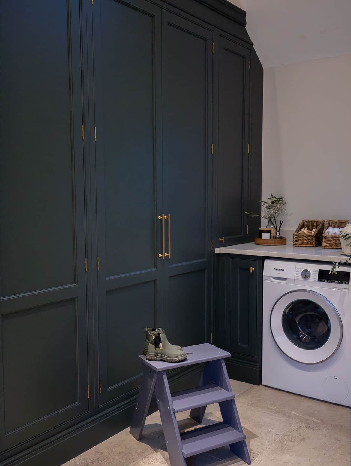 A bespoke bootroom in Downpipe. A view of the floor-to-ceiling bespoke cabinetry with Armac Martin handles. A small purple step stands in front with a pair of wellies while the Siemens washing machine draws the eye towards the bespoke worktop, adorned with incense, two baskets with clothes pegs and hand towels, above it.