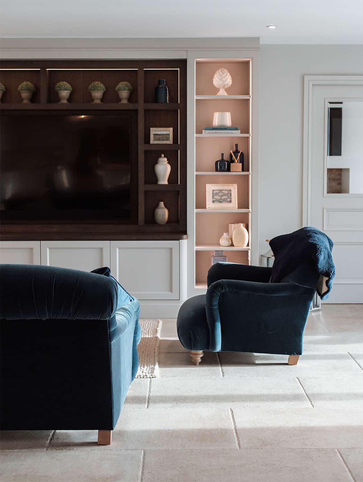A media wall in French Grey. A bespoke media wall overlooks a lounge furnished with off-black sofa and armchair. The flat screen television is positioned in the middle of the wall and surrounded by backlit shelves. The main wall has a selection of ceramic plant pots and jars on the shelves while the columns feature more elaborate jars, urns, and vases. 