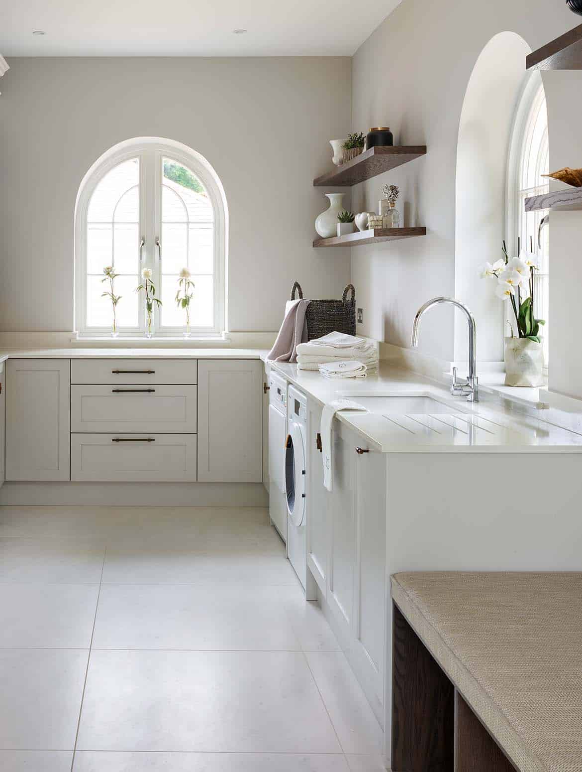 A bespoke bootroom in French Grey. The L-shaped cabinetry feeds around the right side of the view towards the arched window ahead, while the left side has floor-to-ceiling cabinetry that oversees the sink and tap. One shelf lines the right side and two the left of the second arched window. They have a selection of urns, vases and ornaments on them. Beside the sink, a collection of towels and a wicker basket catch the eye. And on the ledge of the far window, three white rose line up in individual vases. A bench stands at the start of the L-shape on the right side of the view. 