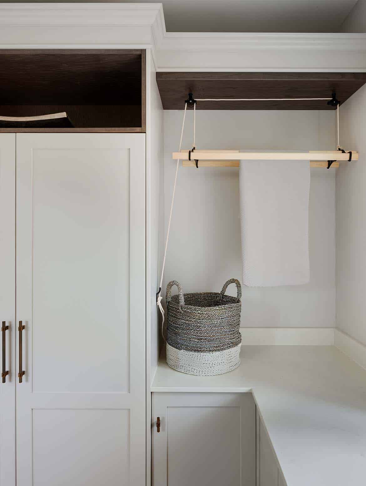 A bespoke bootroom in French Grey. The front on view of the floor-to-ceiling cabinetry to the left marries well with the corner join of the white worktop. The cabinetry continues above the worktop to create a shelf where a small clothes horse hangs down. A single grey towel hangs from it.   