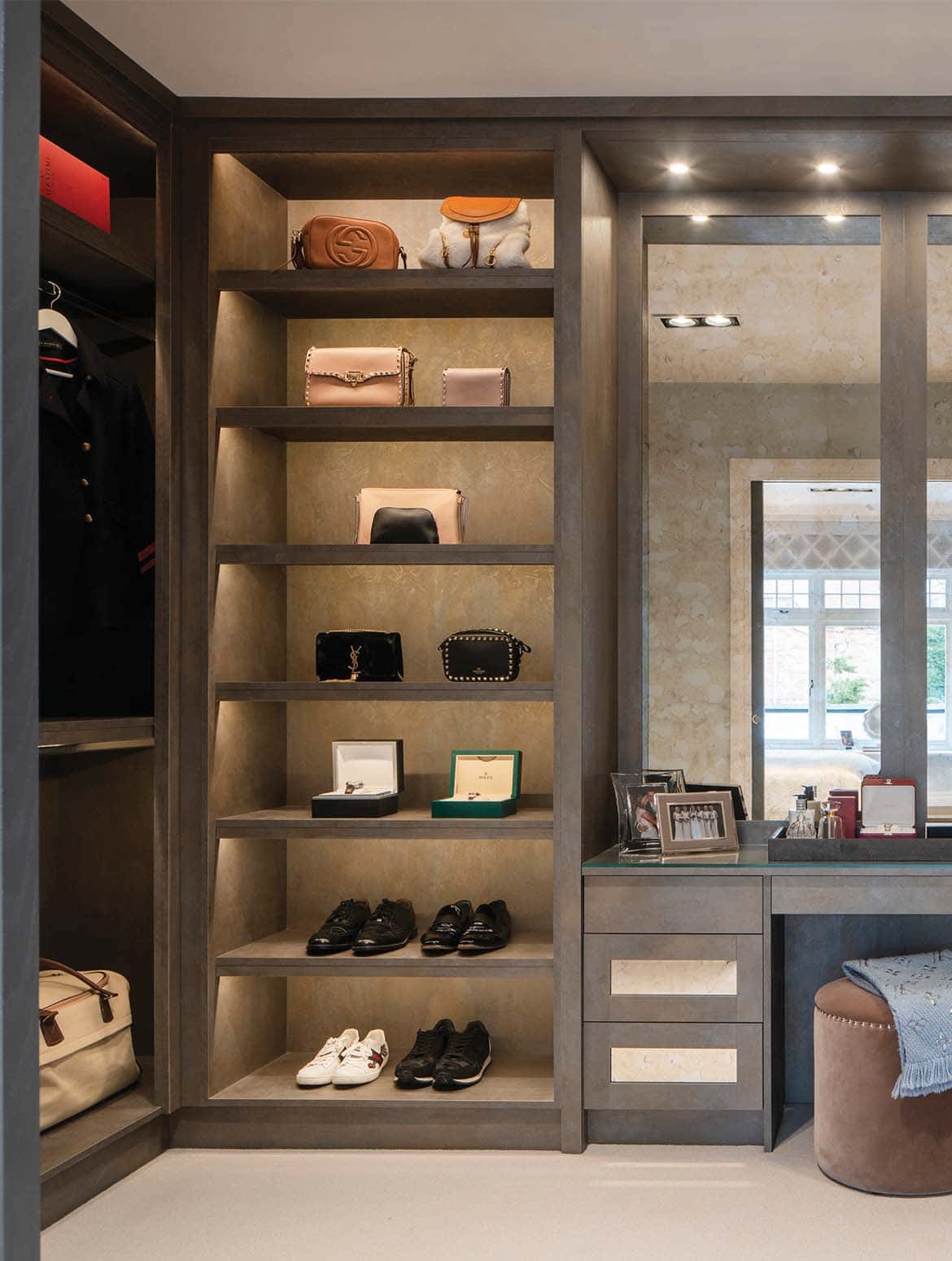 A bespoke dressing room in grey. A corner of bespoke cabinetry has coat and bag storage which leads around to shelving for shoes and bags. This shelving stops to make way for a wide mirror above a vanity built into the cabinetry. 
