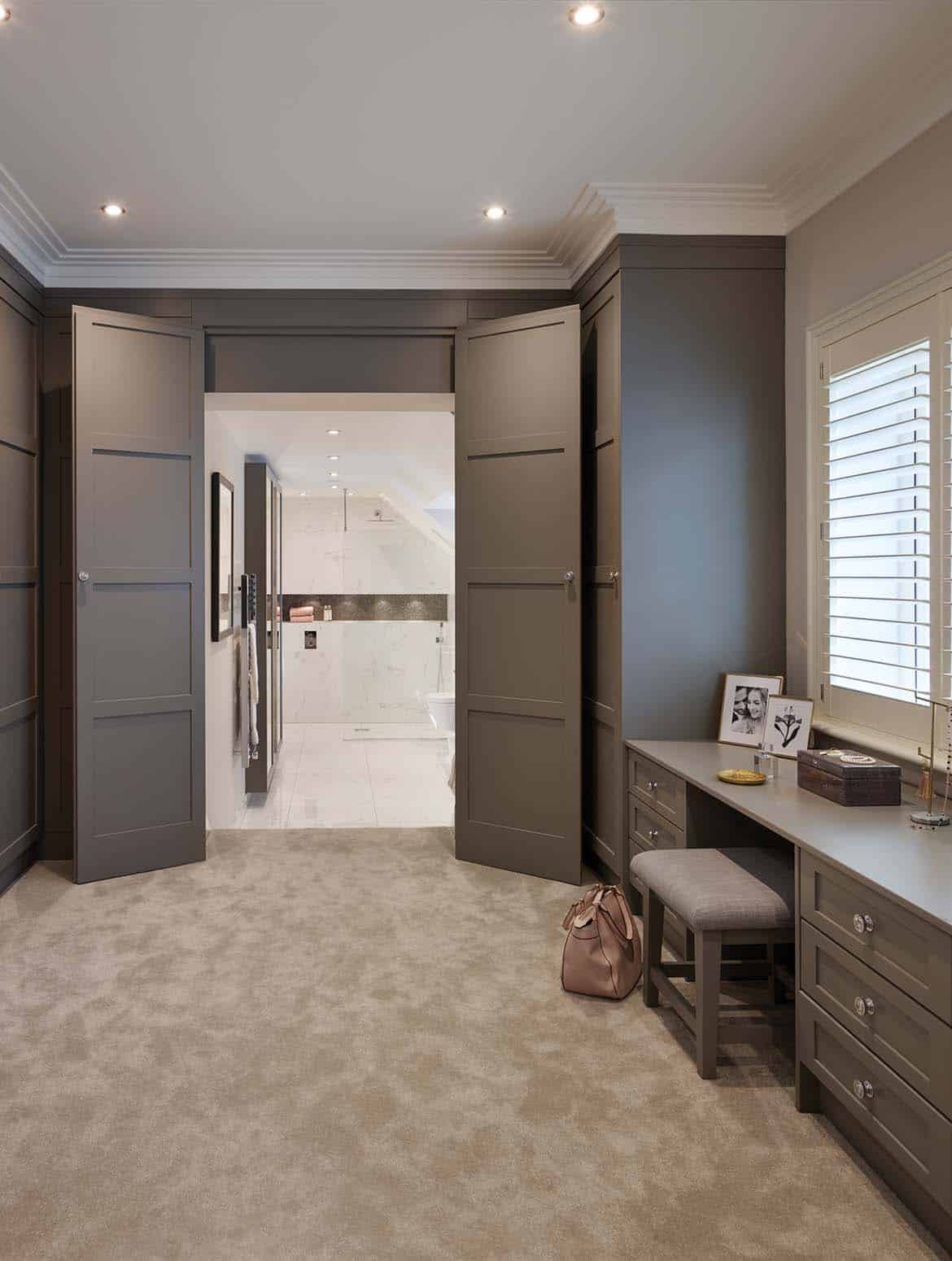 A bespoke dressing room in grey. A plush carpet guides the eye towards the walk in wet room. The bespoke vanity lining the right wall and the bespoke cabinetry along the right helps this view. On the vanity, a couple of frames and small box stand tilted in towards the stool set beneath the vanity. 
