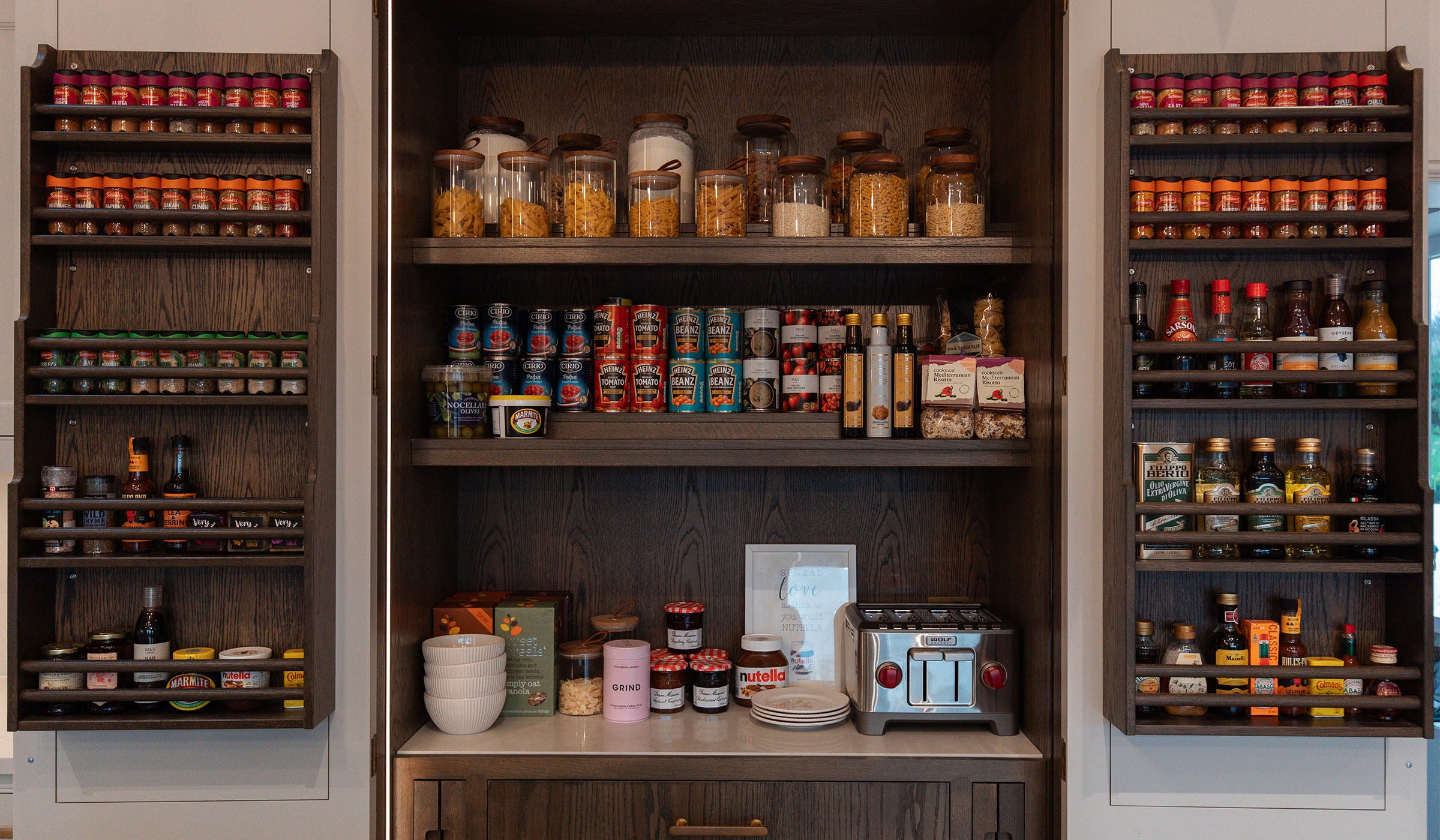 The Pelham Project A traditional shaker kitchen in French Grey. The open pantry doors show two shelves of spices, condiments, and oils. In the main pantry, there is a high shelf with pastas and grains, below that tinned goods, and on the main worktop there are conserves, cereals, and a Wolf toaster.