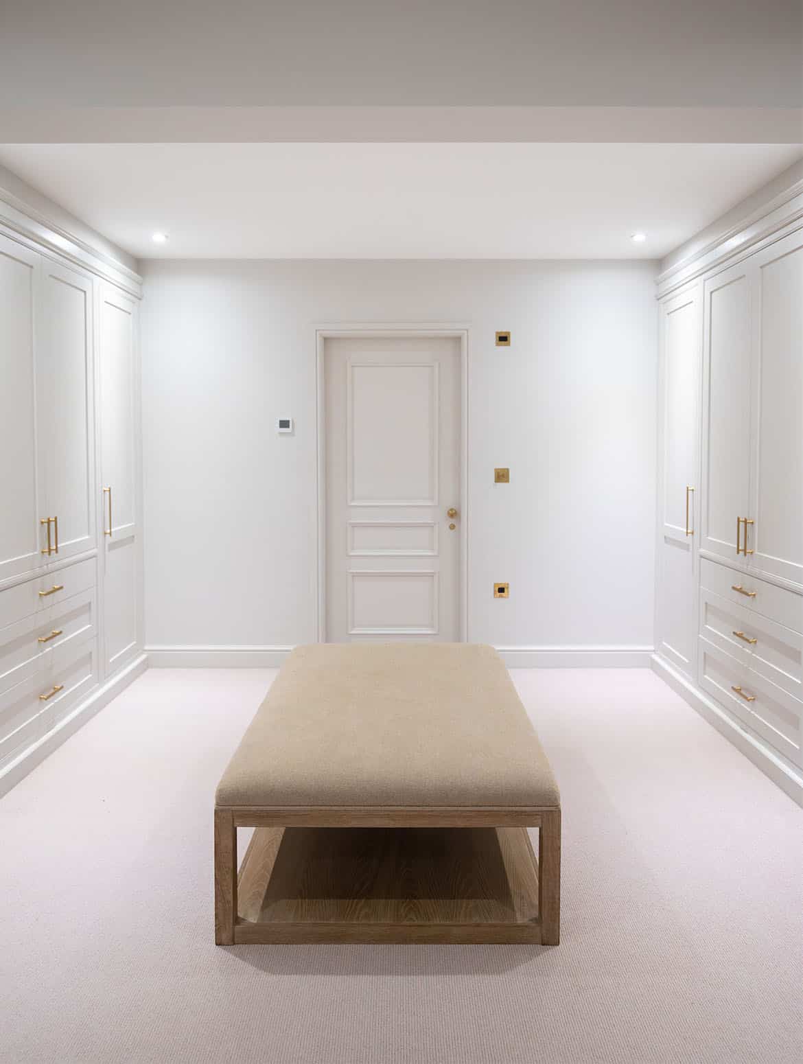 A bespoke dressing room in French Grey. Floor-to-ceiling in frame cabinetry, complete with shaker doors and Corston Architectural handles, stands against either wall. While, an upholstered beige bench stands in the centre of the room ahead of the door-also with Corston Architectural handles. 