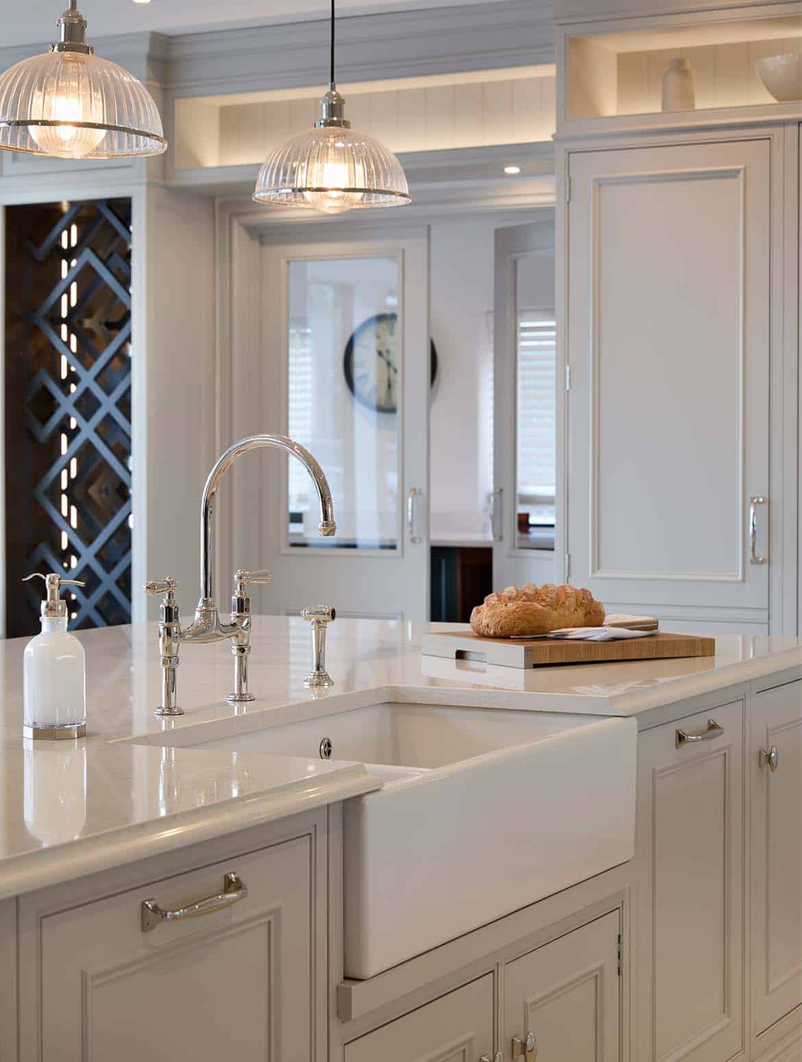 A traditional shaker kitchen in French Grey. The bespoke central island fitted with Snowy Ibiza quartz catches light from three hanging bulbs above. A chopping board with sourdough loaf sits beside the Shaws Belfast sink. Just beyond, floor-to-ceiling cabinetry with Armac Martin satin lacquer finish handles oversees the island. 
