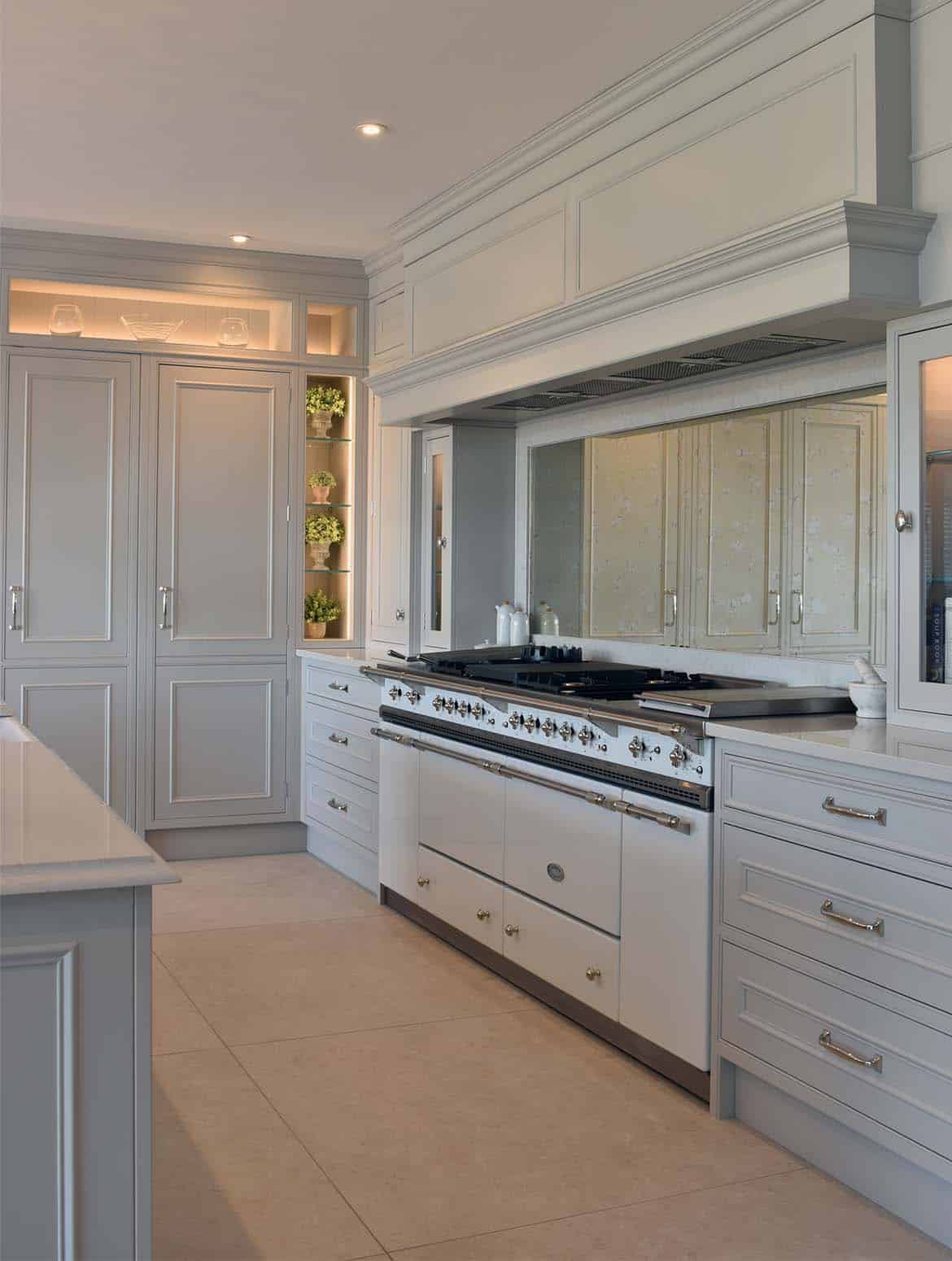 A traditional shaker kitchen in French Grey. The mantel and bespoke cabinetry surrounding a white Lacanche Vezelay range cooker guides the eye to the floor-to-ceiling cabinetry with Armac Martin satin lacquer finish handles. The edge of the bespoke central island fitted with Snowy Ibiza quartz guides the eye from the left side of the frame. 