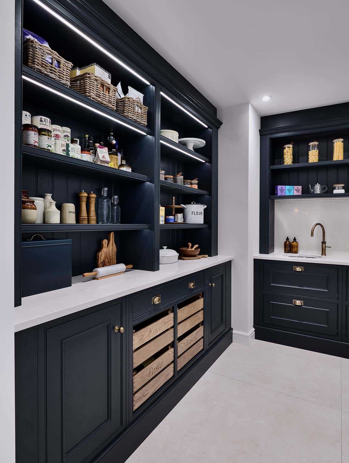 A traditional shaker kitchen in French Grey. The off-black theme of the pantry contrasts the French Grey of the kitchen. Shelves line the wall to the left and the back are adorned with savoury foodstuffs for meals and baking. On the worktop directly on from the open door, a sink fitted with Quooker tap stands next to the edge of the cabinetry. 