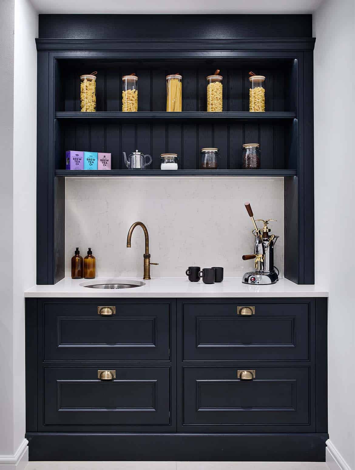 A traditional shaker kitchen in French Grey. The off-black theme of the pantry contrasts the French Grey of the kitchen. On the worktop directly on from the open door, a La Pavoni coffee machine stands next to a sink fitted with Quooker tap. While above, the top shelf holds five jars of different pastas and below the ingredients for a hot beverage are kept.