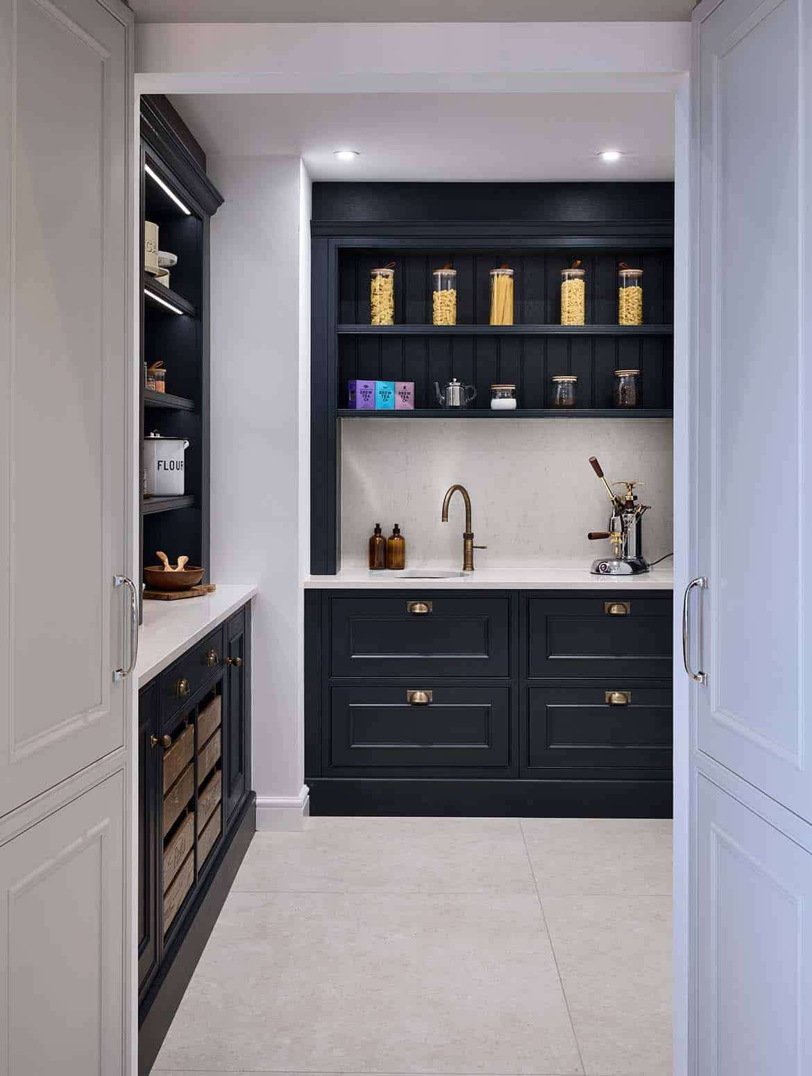 A traditional shaker kitchen in French Grey. The open door to the walk-in pantry has an Armac Martin handle with a satin lacquer. The cabinetry of the pantry can be seen beyond the open door. The off-black theme of the pantry contrasts the French Grey of the kitchen. Shelves line the wall to the left and the back adorned with savoury foodstuffs. On the worktop directly on from the open door, a La Pavoni coffee machine stands next to a sink fitted with Quooker tap. 
