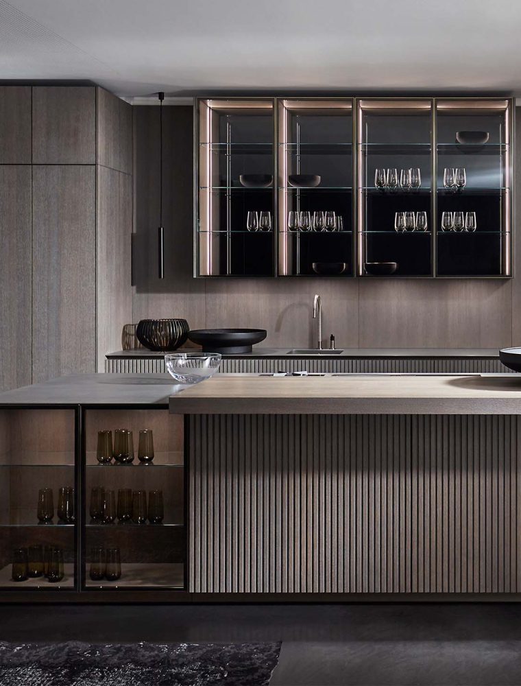 A modern kitchen in smoked wood finish. The floating island stands central to bespoke cabinetry against the back wall. The back wall has push-to-open cabinetry and cupboards with glass fronts and inlaid lighting. Two stools stand below the island’s edge while a modern arm sees to the opposite edge of the island. Set against the left wall, bespoke cabinetry surrounds V-Zug ovens.