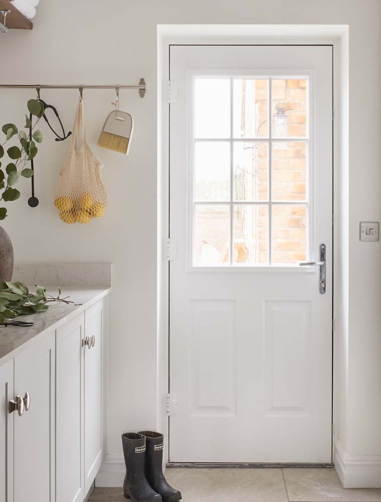 A bespoke bootroom in French Grey. The bespoke cabinetry fitted with handles from Armac Martin’s Bourneville’s collection guides the eye towards the utility door. Nestled within the cabinetry, tumble dryer and washing machine oversee the similarly crafted cupboards and worktop, fitted with Belfast sink. Above the sink, a towel-laden shelf oversees the rail by the door. The rail has a stethoscope, a net of lemons, and a brush hanging from it. A pair of black wellington boots sit by the door and a large ceramic pot stands on the worktop.