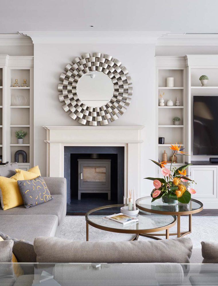 A media wall with bespoke cabinetry in French Grey. Against the far wall, a central fireplace with log burner is set within an Edwardian-style mantelpiece. The left side of the wall has bespoke cabinet with shelves holding an assortment of basks, ceramic urns, vases and decorative plants. The other wall has a television mounted in the middle while the surrounding cabinetry has similar shelving with assortments as well. An L-shaped sofa and two-tier glass coffee table sit in the middle of the lounge.