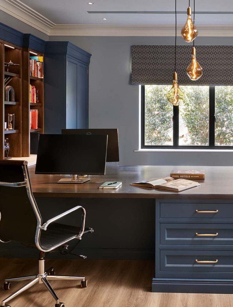 A bespoke home office in off-black. The bespoke cabinetry that runs across the whole left wall houses memorabilia, books, and a mounted television. The TV oversees a desk with space for two people to work opposite each other illustrated by two monitors. The window behind desk lets in a great amount of light giving an ample view from the two comfy chairs set next to large print of the film poster for Spectre (James Bond) on the righthand wall.