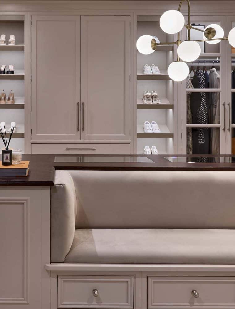 A bespoke modern dressing room interior. The bespoke sofa-cum-storage unit fitted with Englemere and Edwardian panel profiling and Armac Martin handles stands central in the dressing area. One arm of the U-shaped installation has a vase with a bouquet of flowers on it and incense candles on the other. Behind the middle cabinetry, a bespoke floor-to-ceiling wardrobe fitted with two separate rails, two shoe shelves totalling 5, and more space in the middle stands against the far wall. A modern lamp with multiple bulbs within orb-like shading oversees the middle cabinetry.