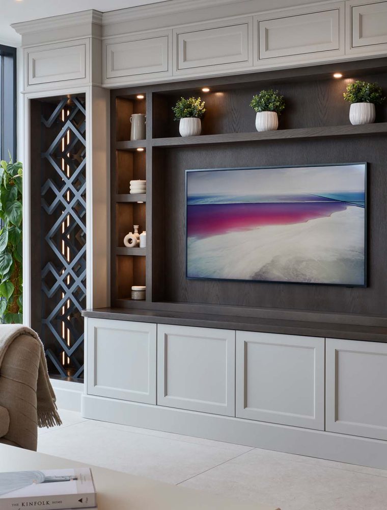 A bespoke media wall in French Grey. The mounted television, focal point the wall, oversees a Tetrad sofa and a coffee table with a ceramic vase on its surface. Surrounding the television, bespoke shelving, set into floor-to-ceiling cabinetry, adds charm through its assortment of ceramic urns and vases, complementing the diagonal woodwork of the main pillars to the cabinet.