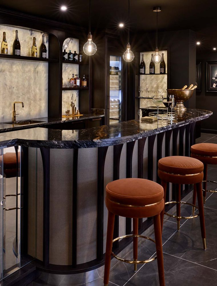 A bespoke bar area in onyx. The well-lit orange velvet upholstered booth catches the eye ahead, while upholstered stools in matching velvet stand before a Orinoco Granite countertop. Beyond the countertop, an onyx backsplash draws attention as well as the gold Quooker tap before it. The overarching cabinetry around the backsplash has a variety of spirits, gins, and champagne. Not to mention, a La Pavoni on the bottom left shelf. 