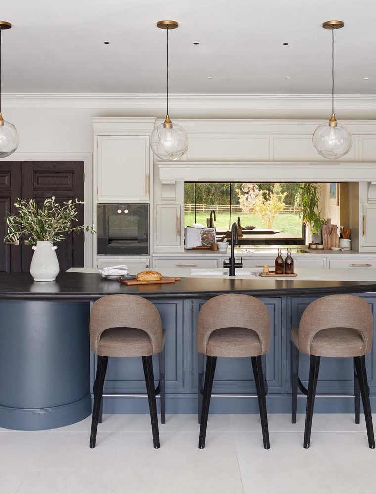 A traditional shaker kitchen project in off-black. A central island with the bespoke cabinetry and a streamlined wood ledge, overseeing three upholstered highchairs, contrasts the white worktop of the remaining island beyond. To its left, bespoke off-black cabinetry oversees the cabinetry opposite that houses Liebherr monolith fridge and freezer. To their right, two Gaggenau ovens are built-in to the middle French Grey cabinetry.