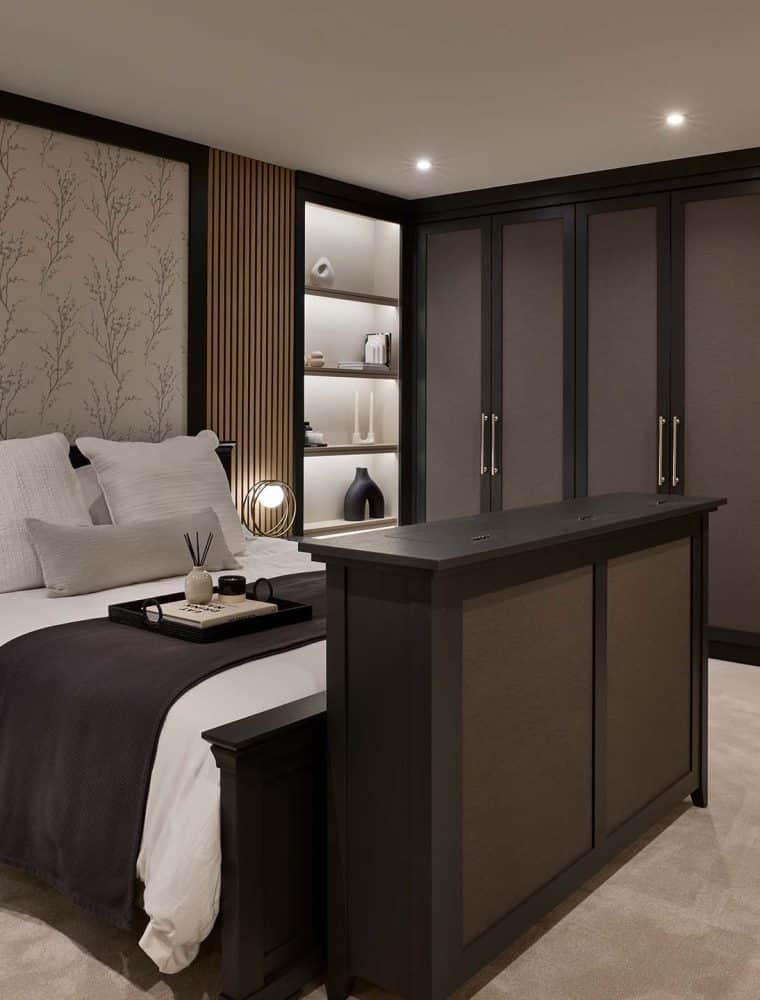 A bespoke modern bedroom interior. The far wall has bespoke floor-to-ceiling wardrobe doors. They lead around to the bespoke wall that frames the bed. Shelving either side the bed board have a mix of ceramic vases and urns. At the foot of the bed, the hinges are just visible of the TV compartment; one that reveals itself from an almost seamless lid. Two bedside tables with circular lamps stand on either side lighting the tray and incense on well-made bed. 