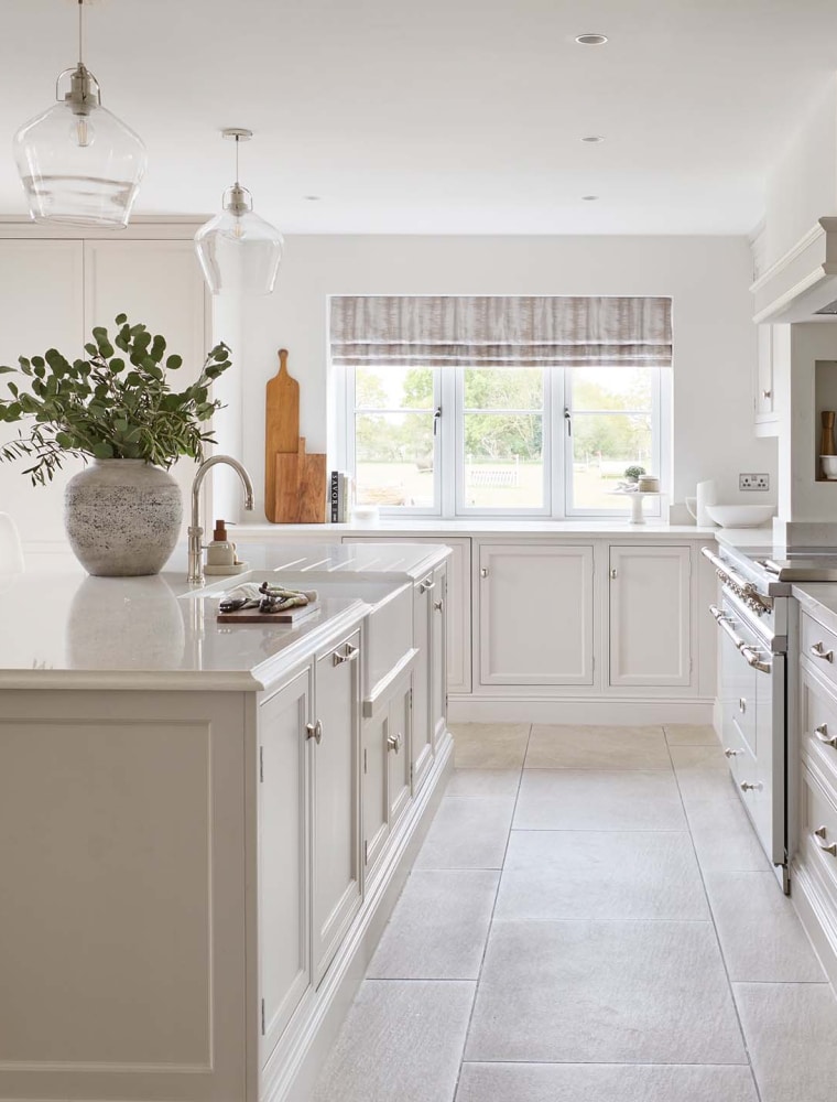 A traditional shaker kitchen in cream. A central island with a Quooker tap and large ceramic vase stands opposite bespoke cabinetry housing a white Lacanche. Large windows at the far end of the kitchen let in a great deal of light over the bespoke worktops.