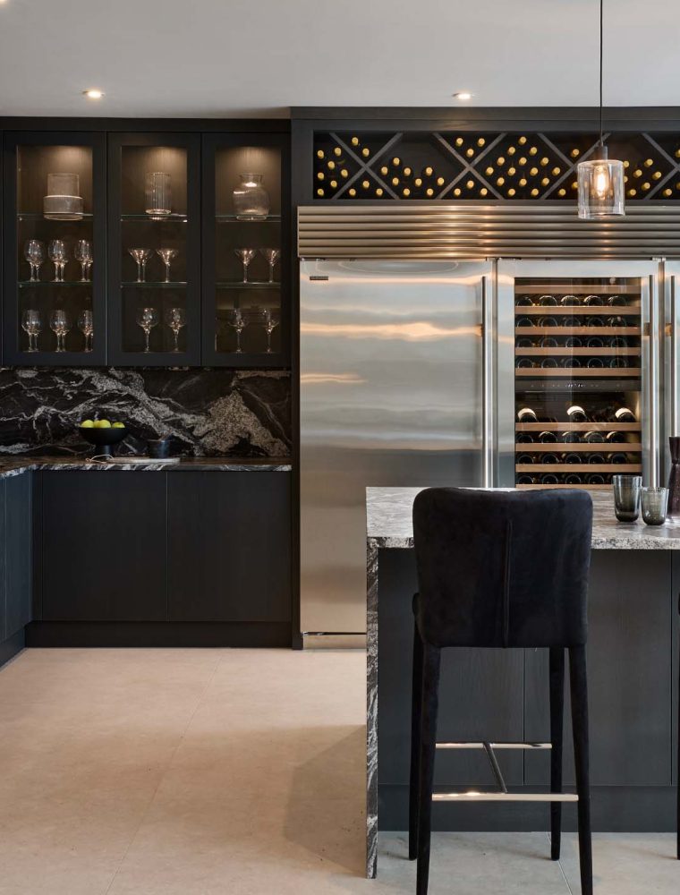 A modern kitchen in Black. The distinct granite central island with waterfall edge overseeing three black upholstered chairs is surrounded by bespoke cabinetry. It begins with the Valencia door to the far right that joins the Sub-Zero refrigeration and worktops beyond. To the far left wall, built in appliances and coffee maker are set into more bespoke panelling.