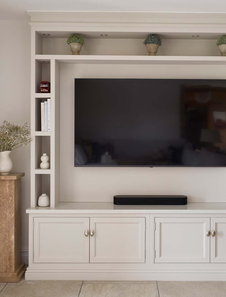 A bespoke media wall in French Grey. The bespoke floor-to-ceiling media wall has Edwardian-styled profiling that stands parallel to the exposed brick wall on its left. The mounted flat screen television stands in the middle of cabinetry crafted with left and right side-shelving including a mantel along the top with three ceramic plants spaced equally along it. Underneath, cupboards with Armac Martin handles sit flush to the cabinetry. A wicker log basket stands to the right of the media wall while a tall plinth with ceramic jug on top stands between the media wall and the exposed brick to the left.