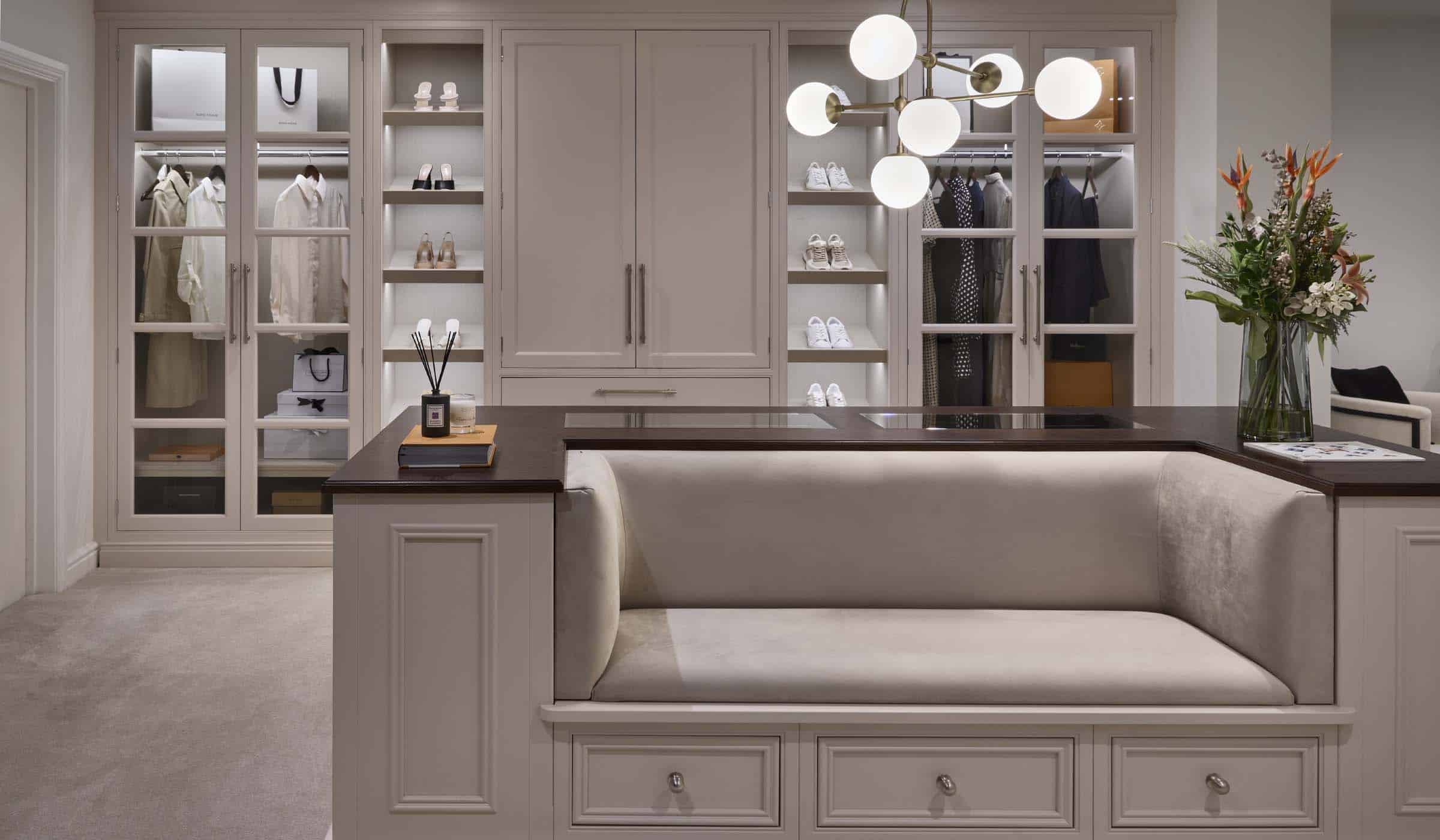 The Shadow Dressing Room Project A bespoke modern dressing room interior. The bespoke sofa-cum-storage unit fitted with Englemere and Edwardian panel profiling and Armac Martin handles stands central in the dressing area. One arm of the U-shaped installation has a vase with a bouquet of flowers on it and incense candles on the other. Behind the middle cabinetry, a bespoke floor-to-ceiling wardrobe fitted with two separate rails, two shoe shelves totalling 5, and more space in the middle stands against the far wall. A modern lamp with multiple bulbs within orb-like shading oversees the middle cabinetry.