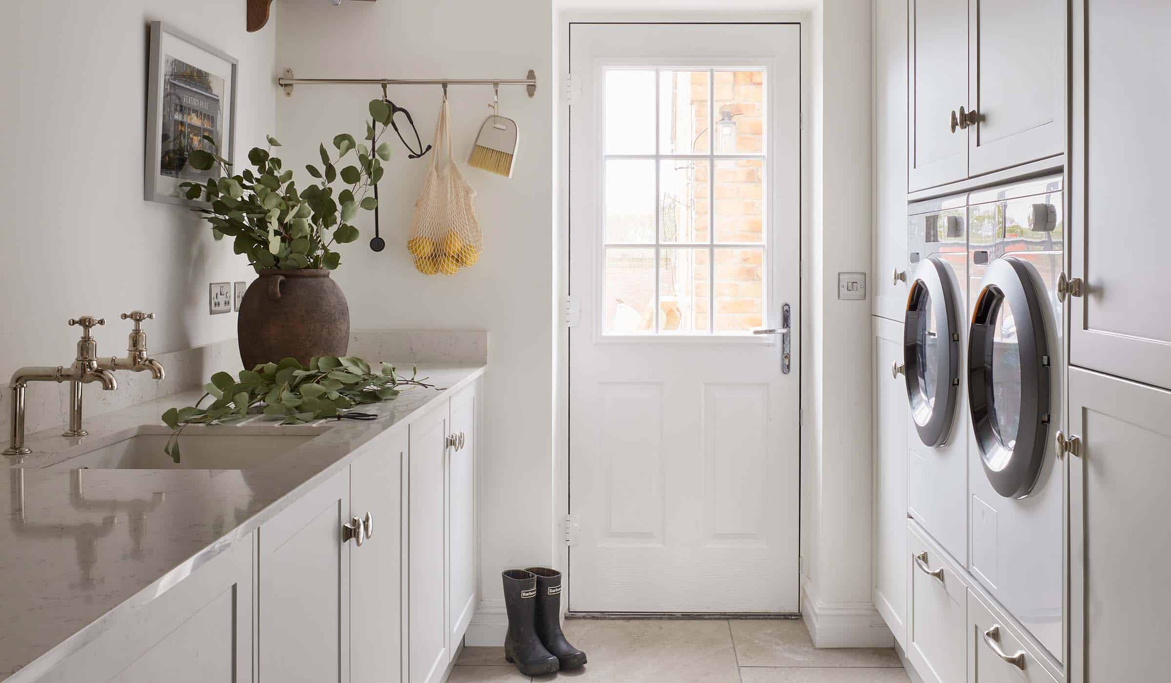A bespoke bootroom in French Grey. The bespoke cabinetry fitted with handles from Armac Martin’s Bourneville’s collection guides the eye towards the utility door. Nestled within the cabinetry, tumble dryer and washing machine oversee the similarly crafted cupboards and worktop, fitted with Belfast sink. Above the sink, a towel-laden shelf oversees the rail by the door. The rail has a stethoscope, a net of lemons, and a brush hanging from it. A pair of black wellington boots sit by the door and a large ceramic pot stands on the worktop.