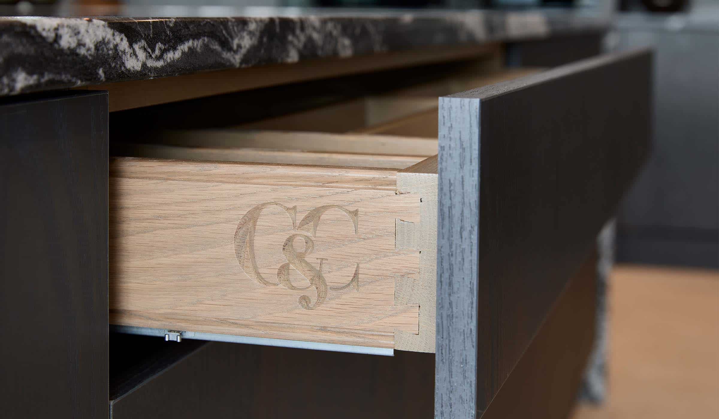 A modern kitchen in Black. The open drawer on the central island has an oak satin finish to it and has the Cooks & Company logo engraved into its edge. While the Valencia panelling on the front ensures a seamless edge.  