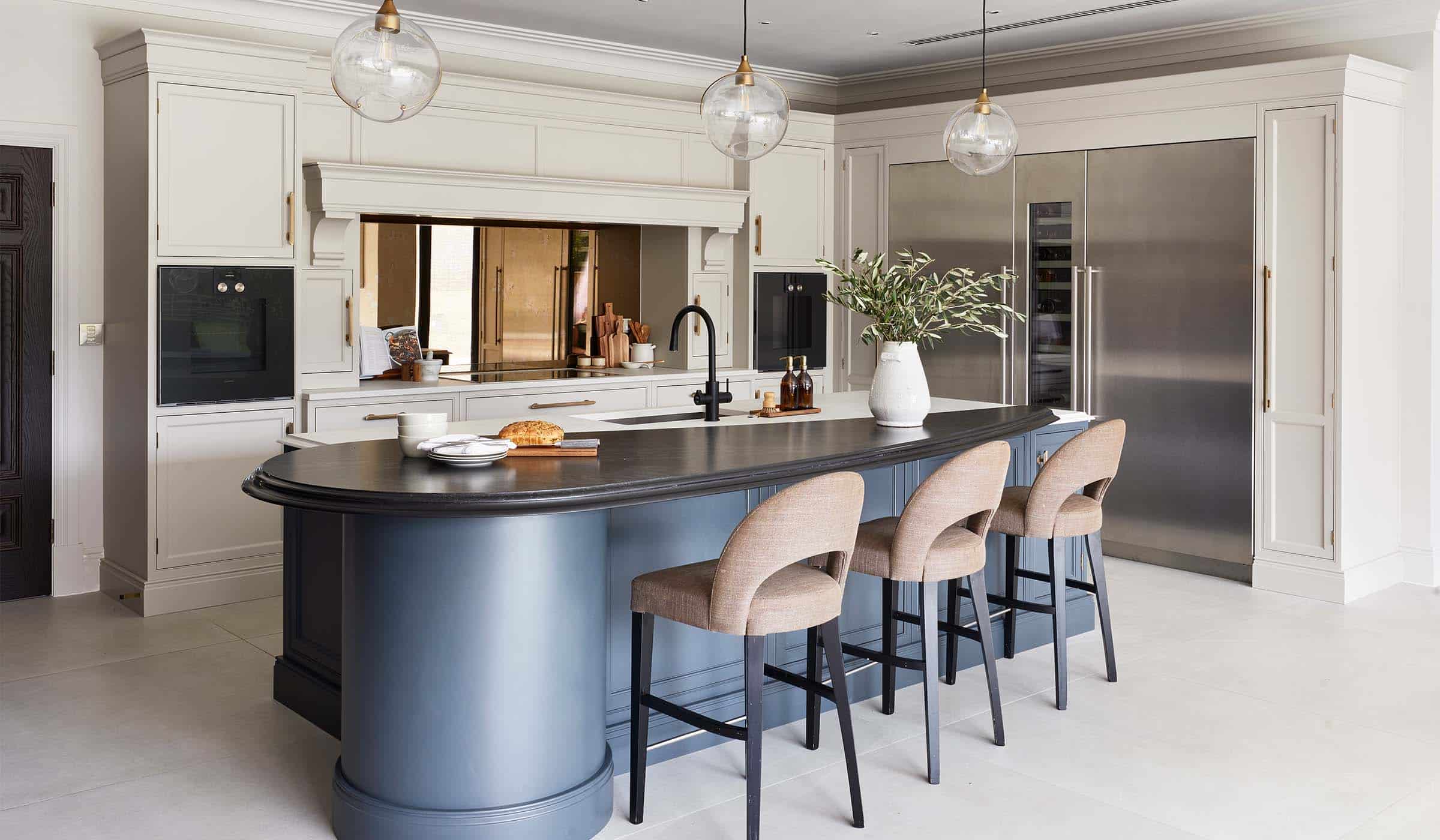 The Tawny Traditional Shaker Kitchen Project A traditional shaker kitchen project in off-black. A central island with the bespoke cabinetry and a streamlined wood ledge overseeing three upholstered highchairs contrast the white worktop of the remaining island beyond. To its right, two Liebherr monoliths are set within French Grey bespoke cabinets and against the back wall, two Gaggenau ovens are built-in to the middle French Grey cabinetry. The mantel stands proud to the surrounding cabinetry.