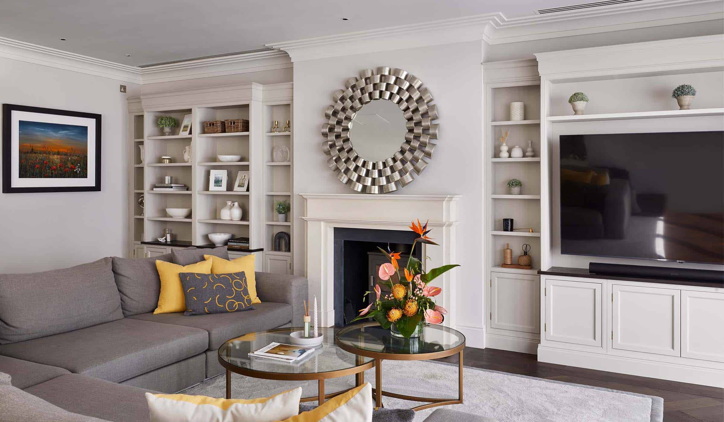 A media wall with bespoke cabinetry in French Grey. Against the far wall, a central fireplace with log burner is set within the Edwardian-style mantelpiece. The left side of the wall has bespoke cabinet with shelves holding an assortment of baskets, ceramic urns, vases and decorative plants. The other wall has a television mounted in the middle while the surrounding cabinetry has similar shelving with assortments as well. An L-shaped sofa and two-tier glass coffee table sit in the middle of the lounge.  