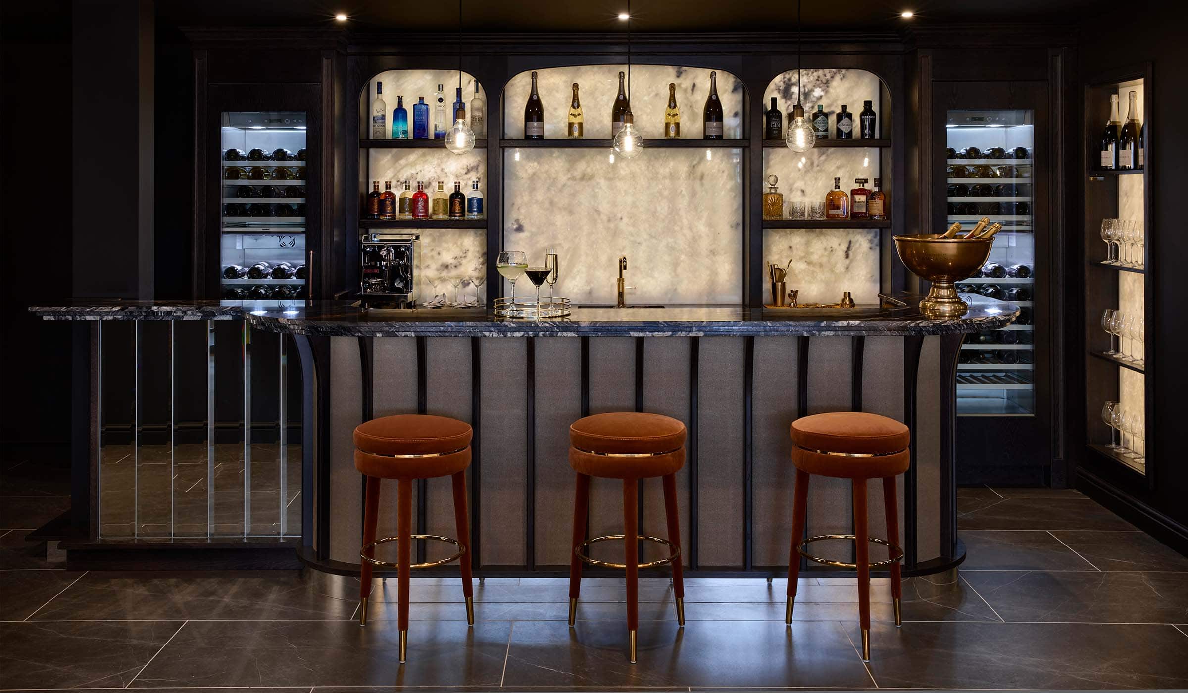 The Bingham Bar Project A bespoke bar area in onyx. The upholstered stools in orange velvet catch the eye stood before a Orinoco Granite countertop. Beyond the countertop, an onyx backsplash draws attention as well as the gold Quooker tap before it. The overarching cabinetry around the backsplash has a variety of spirits, gins, and champagne. Not to mention, a La Pavoni on the bottom left shelf. On the countertop itself a tray with three drinks: champagne, gin and tonic, and a cocktail are visible.