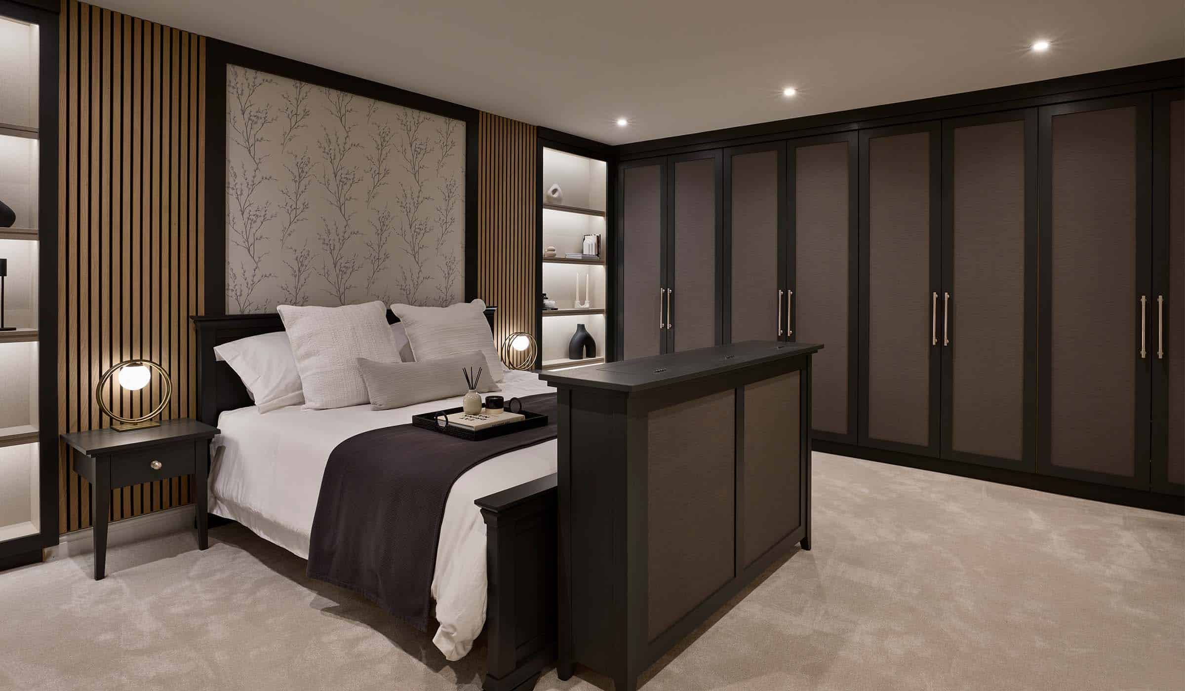 The Willow Dressing Room Project A bespoke modern bedroom interior. The far wall has bespoke floor-to-ceiling wardrobe doors. They lead around to the bespoke wall that frames the bed. Shelving either side the bed board have a mix of ceramic vases and urns. At the foot of the bed, the hinges are just visible of the TV compartment; one that reveals itself from an almost seamless lid. Two bedside tables with circular lamps stand on either side lighting the tray and incense on well-made bed.