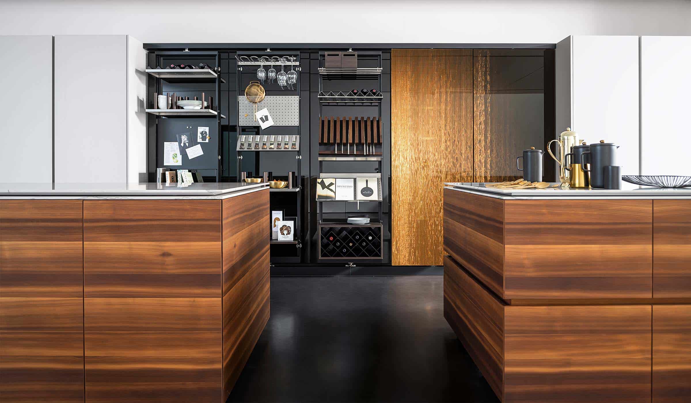 The Larch Modern Kitchen Project A modern kitchen with quartz-top islands. The dual central kitchen islands stand proud to the sleek cabinetry against the far wall which encloses built-in ovens on one side and culinary items in the middle. The open door reveals an ergonomic selection of shelves and racks that have items ranging from wine glasses to a sieve.