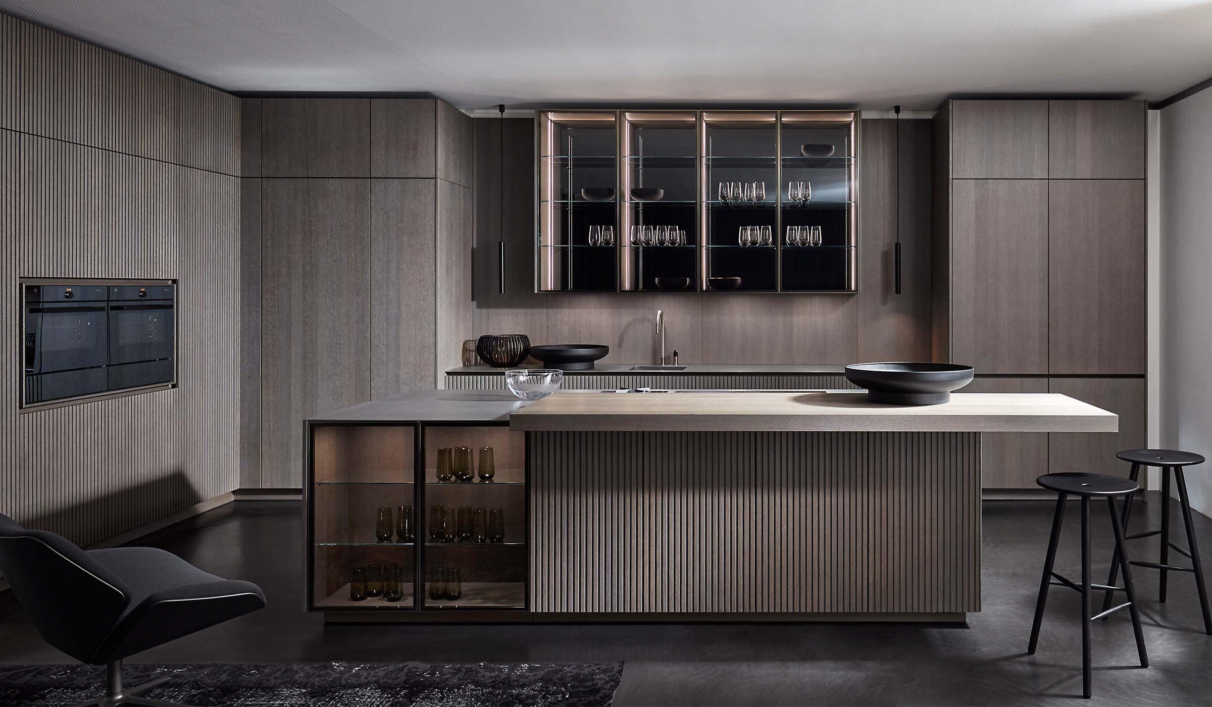 A modern kitchen in smoked wood finish. The floating island stands central to bespoke cabinetry against the back wall. The back wall has push-to-open cabinetry and cupboards with glass fronts and inlaid lighting. Two stools stand below the island’s edge while a modern arm sees to the opposite edge of the island. Set against the left wall, bespoke cabinetry surrounds V-Zug ovens.