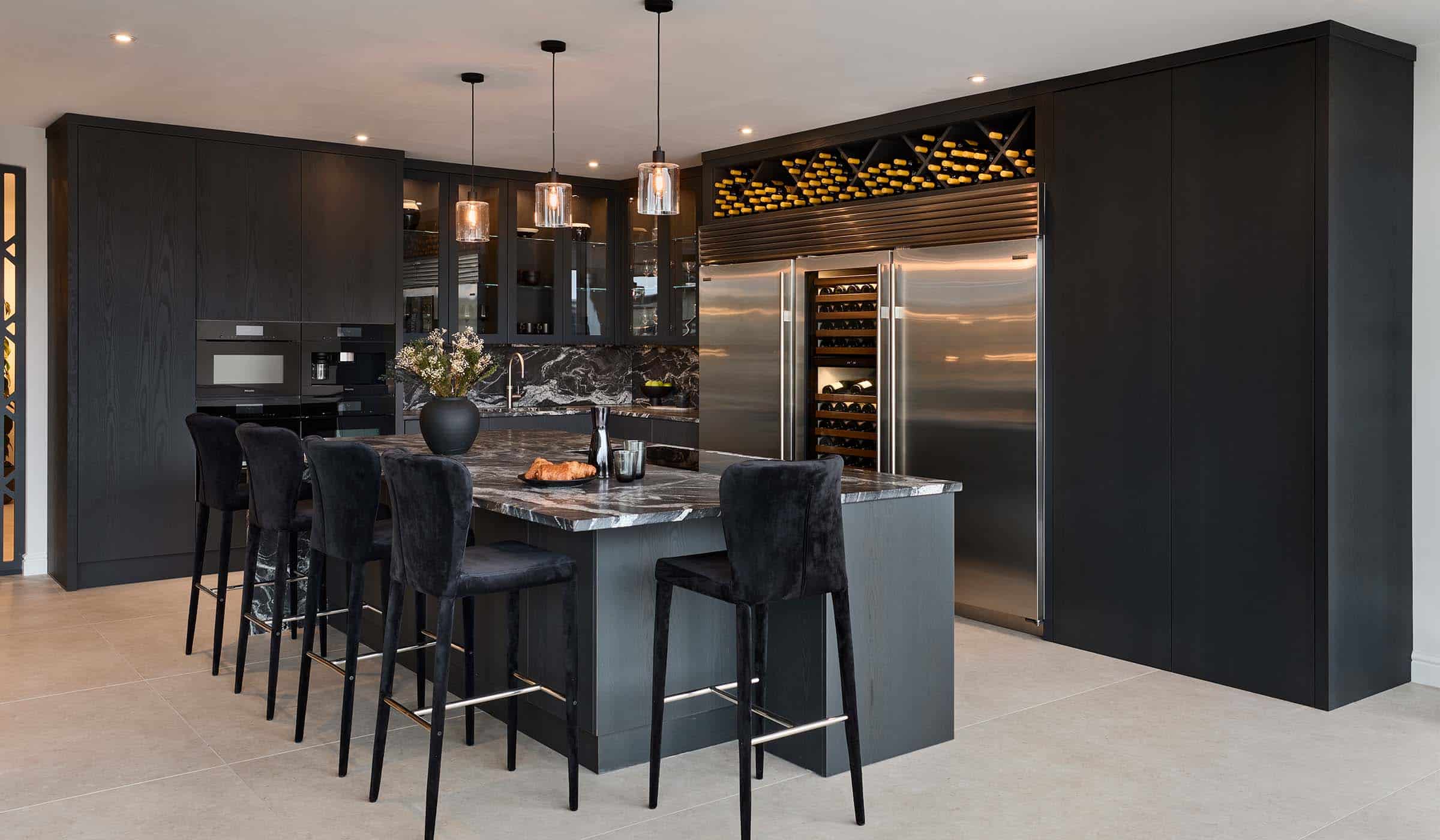 A modern kitchen in Black. The distinct granite central island with waterfall edge overseeing three black upholstered chairs is surrounded by bespoke cabinetry. It begins with the Valencia door to the far right that alludes to the double pantry behind and joins the Sub-Zero refrigeration and worktops beyond. To the far left wall, built in appliances and coffee maker are set into more bespoke panelling. 