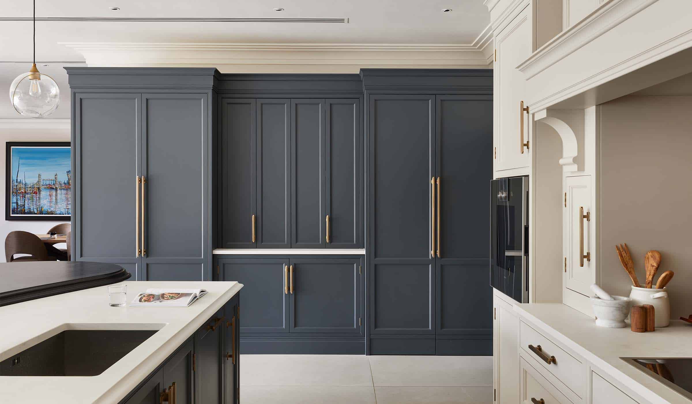 The Tawny Traditional Shaker Kitchen Project A traditional shaker kitchen project in off-black. The edge of the central island with bespoke cabinetry and a streamlined wood ledge contrasts the white worktop of the remaining island beyond. Beyond the island, the bespoke cabinetry of a hidden pantry and other cupboards forms the background. To its edge, there is a small raised table with two high chairs and a framed portrait of London Bridge on the wall behind.