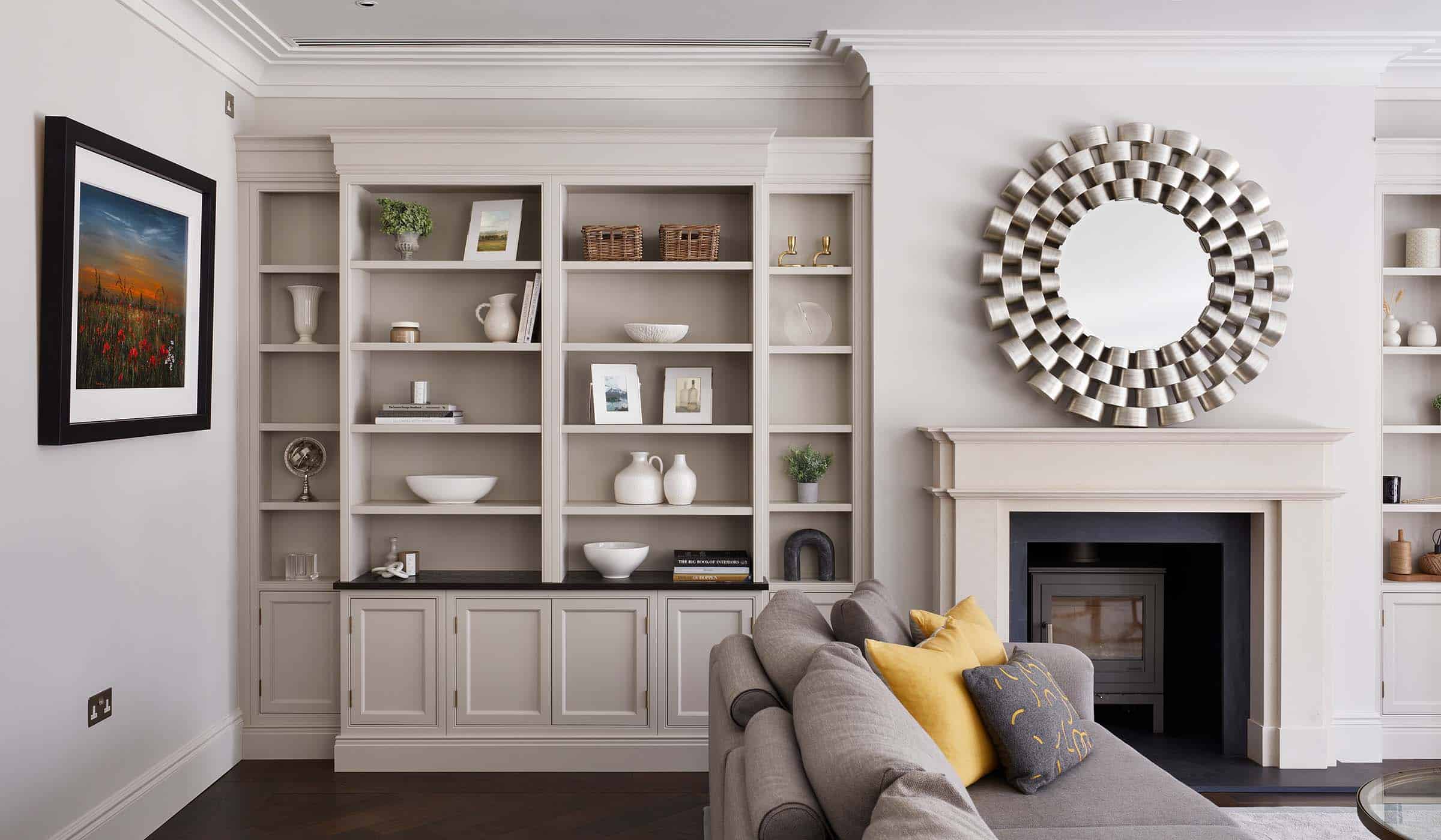 A media wall with bespoke cabinetry in French Grey. Against the far wall, a central fireplace with log burner is set within the Edwardian-style mantelpiece. The left side of the wall has bespoke cabinet with shelves holding an assortment of basks, ceramic urns, vases and decorative plants. An L-shaped sofa and two-tier glass coffee table sit in the middle of the lounge. On the right-angle from the left cabinetry a framed print of a poppy field oversees the lounge beyond.  