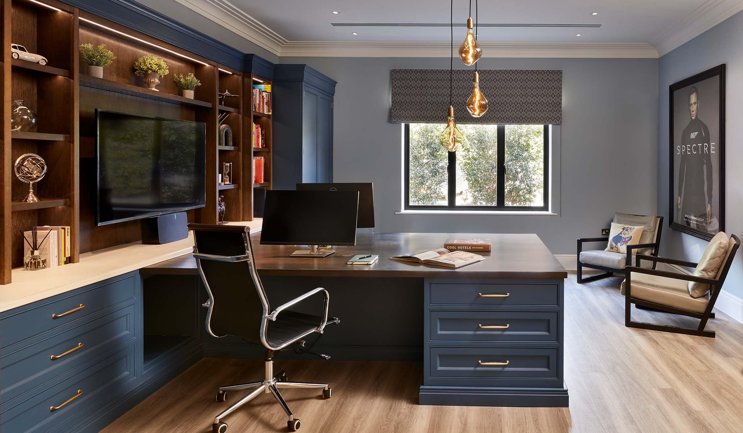 The Tawny Home Office Project A bespoke home office in off-black. The bespoke cabinetry that runs across the whole left wall houses memorabilia, books, and a mounted television. The TV oversees a desk with space for two people to work opposite each other illustrated by two monitors. The window behind desk lets in a great amount of light giving an ample view from the two comfy chairs set next to large print of the film poster for Spectre (James Bond).