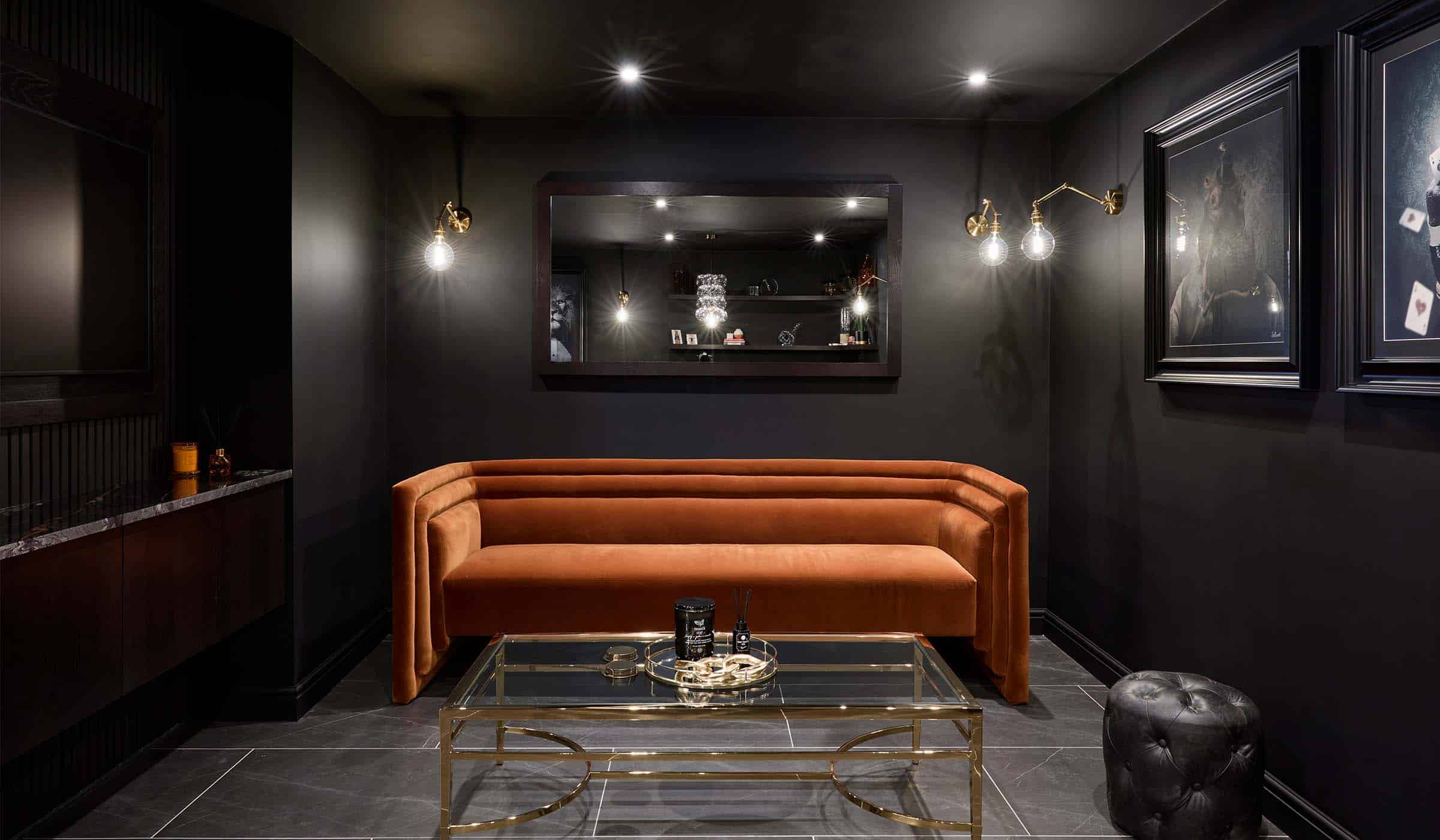 The Bingham Bar Project A bespoke bar area in onyx. The orange velvet sofa set against the back wall catches the light from exposed bulbs above. A glass coffee table with gold edges and leg stands before it along with a black velvet footstool. On the back wall, a mirror reflects the chandelier ahead of the sofa. The flat screen television set into its own media wall is just visible on the left wall along with the artwork opposite revealing slighting a hippopotamus in a party hat.