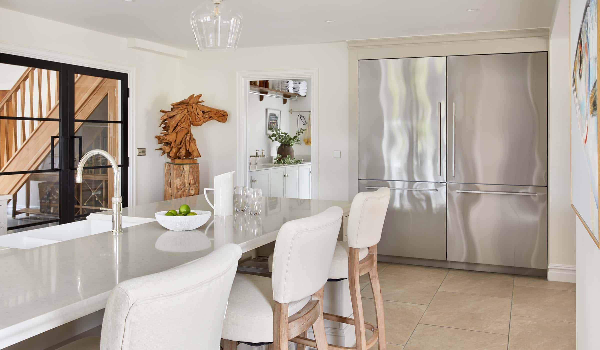 The Fox Traditional Shaker Kitchen Project A traditional shaker kitchen in French Grey. The central island fitted with a Quooker tap has a bowl with apples and a ceramic water jugs with glasses beside it. Beyond the island, there is the doorway to a utility room and next to it is an American-style Fisher and Paykel fridge-freezer. In the other other corner beside the utility doorway, there is a wooden horse sculpture.