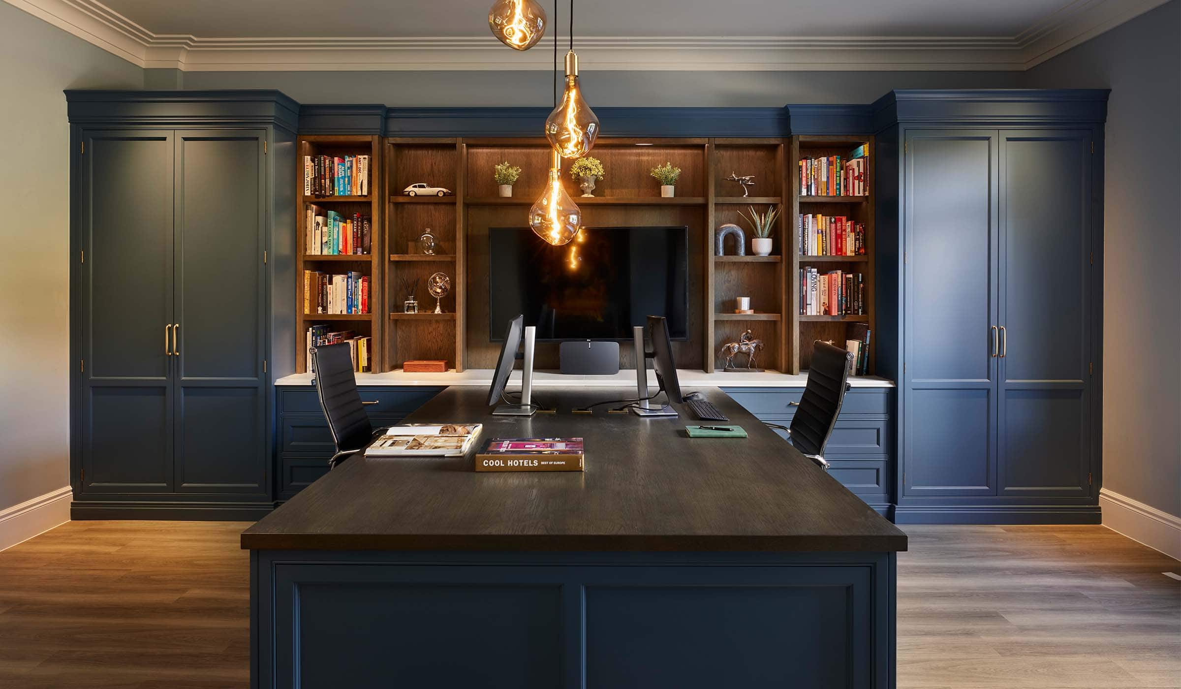 The Tawny Home Office Project A bespoke home office in off-black. The bespoke cabinetry that runs across the whole left wall houses memorabilia, books, and a mounted television. The TV oversees a desk with space for two people to work opposite each other illustrated by two monitors and chairs. Warm hanging lighting oversees the desk while the burnished brass handles of the two cabinets at either end of the cabinets stand out as well.