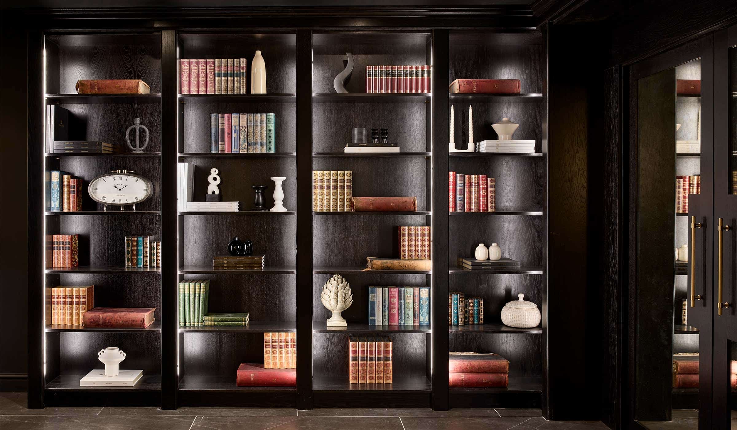 The Bingham Bar Project A bespoke bar area in onyx. The four off-black library shelves hold an assortment of ornaments, books, and tomes on their side. The shelves have a ghostly quality with the inbuilt light around their periphery.